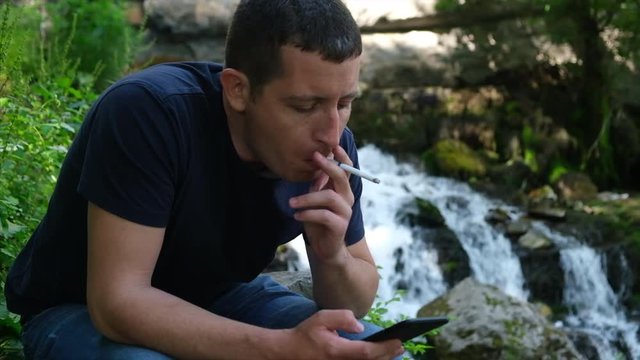 Young alone man browsing social media on a phone and smoking cigarette near a waterfall.