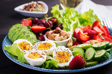 Plate with a paleo diet food. Boiled eggs, avocado, cucumber, nuts, cherry and strawberries. Paleo breakfast. - obrazy, fototapety, plakaty