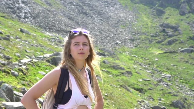 Girl Looks out into the mountains as she contemplates and thinks, standing in the sun surrounded by rocks and green grass in slow motion as her hair blows in the wind