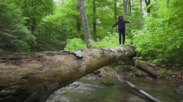 A girl balances on a log bridge as she crosses a small stream while hiking.