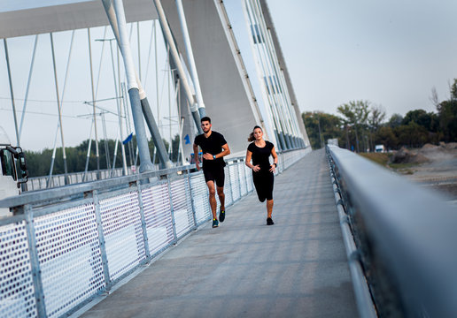 Young Couple In Black Sports Outfit Doing Morning Workout Outdoors. Young Man And Woman Running On Bridge.