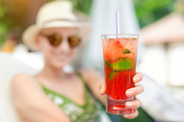 Fototapeta premium Young beautiful happy woman holding in hand and give glass with fresh cold strawberry non-alcoholic mojito cocktail enjoying summer vacation at poolside resort on hot sunny day.Refreshment cold drink