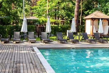 Poolside with clear blue water and loungers under umbrellas on summer day heat. Swimming pool at resort in green coniferous pine forest. Weekend destination place
