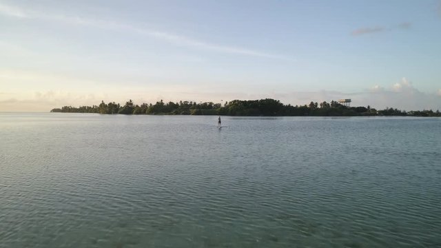 Flying over Maldives ocean, Man Stand Up Paddleboarding away from tropical island in distance
