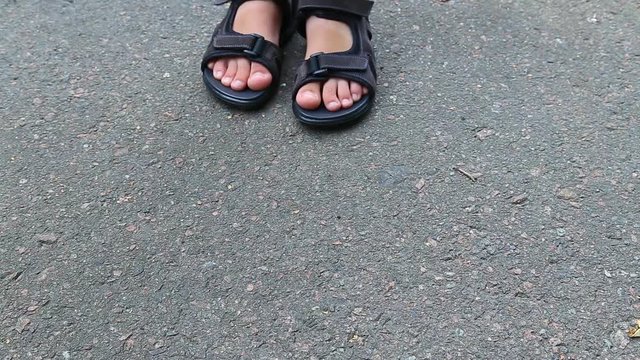 Closeup top view of small child's legs wearing black leather sandals isolated on grey pavement background. Real time full hd video footage.