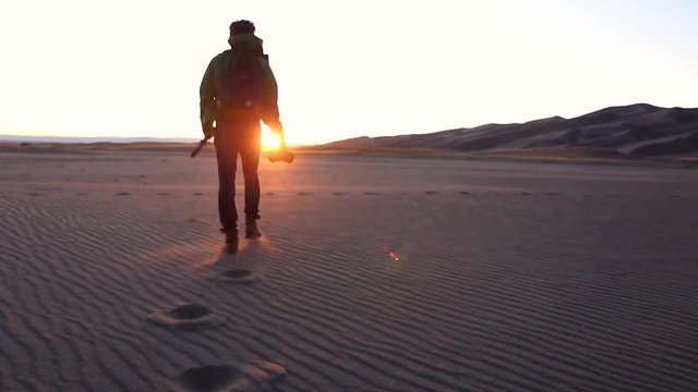 Low angle medium shot of a man with a camera walking across the desert into the sunset