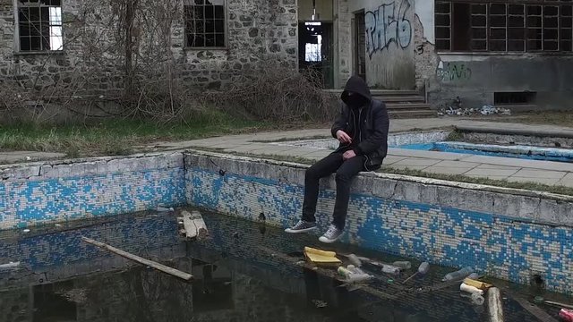 WIDE SLOW MOTION PAN RIGHT: Young male thug causing mischief, wearing black hood sits by abandoned swimming pool and throws a stone into the pool