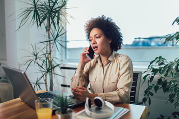 Smiling young African female entrepreneur sitting at a desk in her home office working online with...
