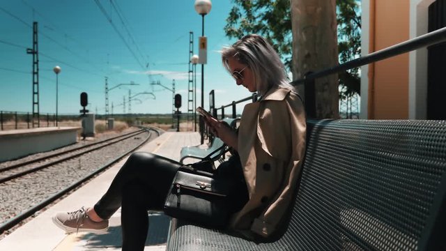 Young beautiful tourist woman checking her smartphone while waiting for the commuter train, wearing sunglasses on a bright sunny day, 4k side view