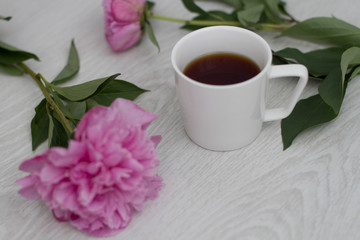 A white Cup of black coffee stands on a white wooden background next to pink peonies