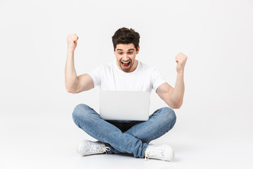 Excited young man posing isolated over white wall using laptop computer sitting on floor make...