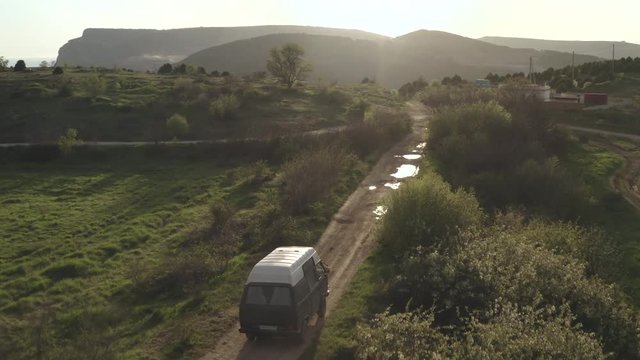 Tourist van car rides winding rural road among bushes and fields. Sandy road passes through hilly area with mountains background. Top view from drone. Travelling by car