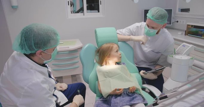 Little caucasian girl in dental chair talking to the dentist and his assistant.