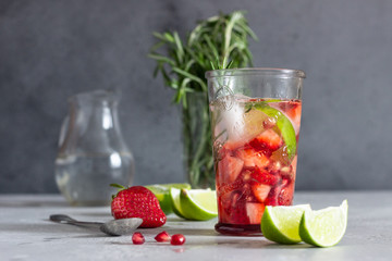 Homemade strawberry lemonade in glass with fresh strawberries, rosemary, pomegranate and lime over light grey stone table. Refreshing summer drink. Cocktail bar background concept. Copy space.