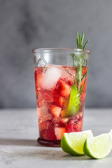 Homemade strawberry lemonade in glass with fresh strawberries, rosemary, pomegranate and lime over light grey stone table. Refreshing summer drink. Cocktail bar background concept. Copy space.