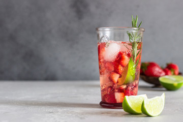 Homemade strawberry lemonade in glass with fresh strawberries, rosemary, pomegranate and lime over light grey stone table. Refreshing summer drink. Cocktail bar background concept. Copy space.
