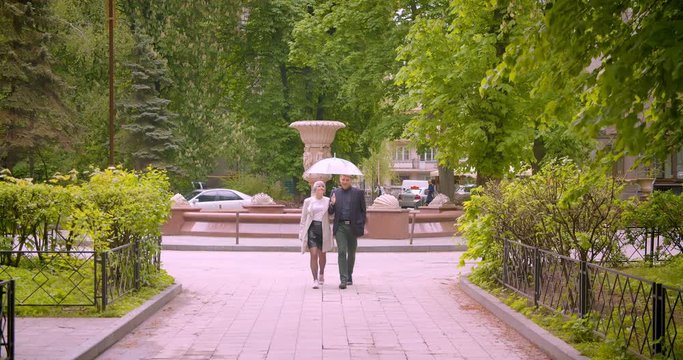 Closeup shoot of young beautiful hipster couple holding an umbrella walking together towards camera being happy in the park outdoors outdoors