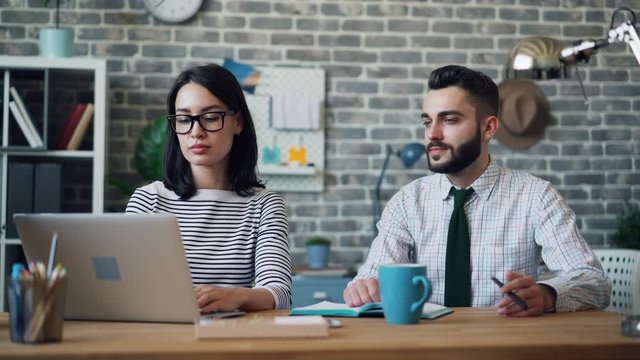 Cinemagraph loop of man moving pen in hand working in office with young woman sitting at desk looking at laptop screen. People, cooperation and computers concept.