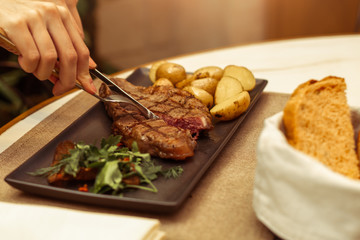 Focus on the meat where the fork and knife. Beautiful female hands in a restaurant at the table cut beef striploin steak.