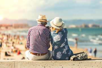 Mature couple of retired lovers enjoying retirement on the beach facing the sea with mobile cell...