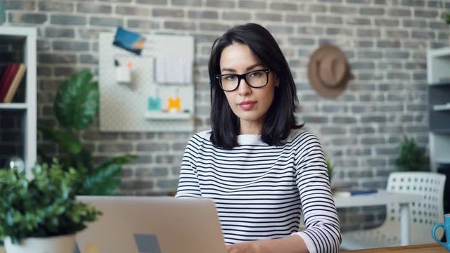 Portrait of beautiful busineswoman in glasses working with laptop then smiling looking at camera sitting at desk busy with work. People and occupation concept.