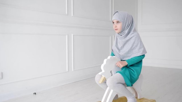 Muslim teen 9 year girl in grey hijab and blue dress is playing riding on toy horse rocking chair in her white modern room.