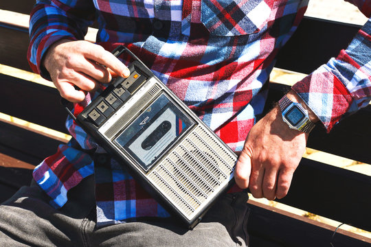 Hipster fancy man in plaid shirt holding in hand and sitting on bench with retro vintage old 80s , 90s style portable radio audio cassette recorder player boombox with audio tape cassette inside 