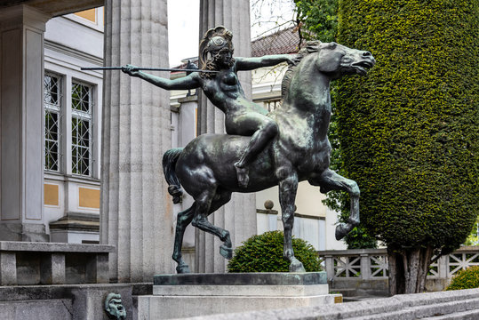 Germany, Munich, Bogenhausen: Equestrian statue with Amazone on horse in front of famous Villa Stuck Museum in the city center of the Bavarian capital with pillars and green bushes.