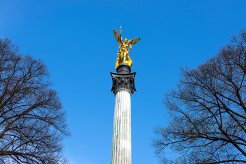 Germany, Munich, Bogenhausen: Steeple of the Peace Column with famous golden Angel of Peace statue (Friedensengel) at the top in the city center of the Bavarian capital with trees and blue sky.