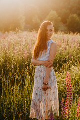 Beautiful Russian young woman in summer dress with long hair standing in a blooming field smiling and resting