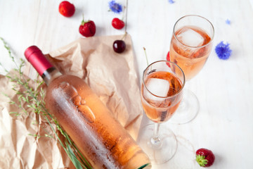 Glasses of rozvogo wine with ice, a bottle of wine, strawberries, cherries and flowers on a white wooden background