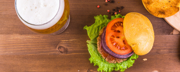 Tasty homemade burger with onions, tomatoes, cutlet and leaf of lettuce and a glass of beer on a wooden table, top view, copy space