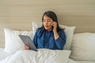 Young woman sitting on bed and using phone and tablet