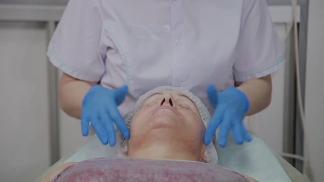 Professional cosmetologist applies protective cream to the elderly woman in the beauty parlor.