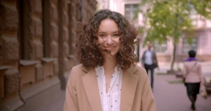 Closeup shoot of young long haired curly caucasian female student smiling happily walking towards camera outdoors on the street with blurred focus