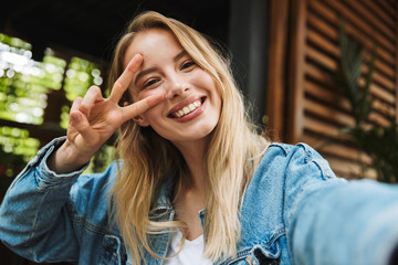 Happy young pretty blonde woman in cafe take a selfie by camera showing peace.