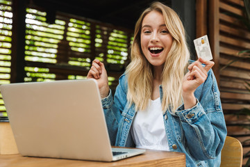 Happy young pretty blonde woman in cafe using laptop computer holding credit card.