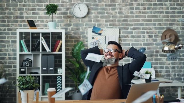 Slow motion of lots of money falling on handsome middle-aged guy in office sitting at desk alone enjoying wealth. Happiness, rich youth and financial luck concept.