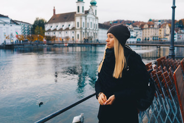 Young blonde tourist girl in warm hat and coat with backpack walking at cold autumn in Europe city enjoying her travel in Lucerne Switzerland - obrazy, fototapety, plakaty