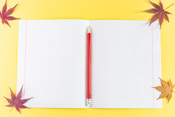 Notebook in a cage, a red pencil with an eraser and autumn maple leaves on a bright yellow background. The concept of the day of knowledge, the first of September. Beginning of the school year. 