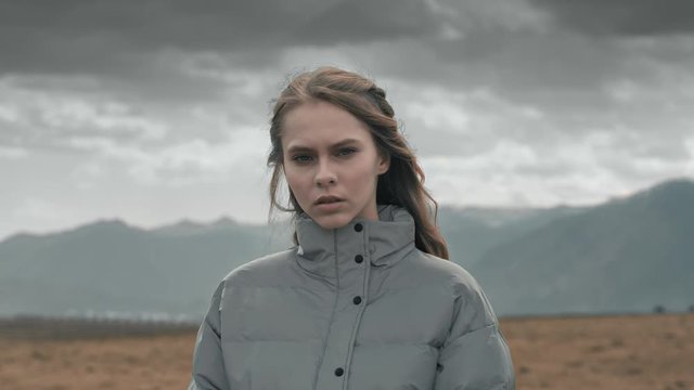 Close-up portrait: a beautiful young girl with long hair in a gray jacket stands in the open nature against the mountains, looking at the camera, hair develops wind. Grey cloudy light, slow motion, 4K