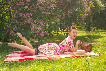 sweetheart charming pinup girl in a summer dress on a checkered blanket in the Park near the bushes of lilac enjoys life and leisure alone. pretty happy caucasian woman on the grass.