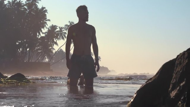 handsome man in shorts silhouette stands in shallow water against palms with brown rock on foreground slow motion. Concept wellness lifestyle