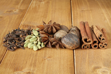 Pile of dry spice cloves, pile of dry scattered unrefined cardamom seeds, bunch of dry anise spice, dried nutmeg fruits, dry cinnamon sticks, lying on wooden boards