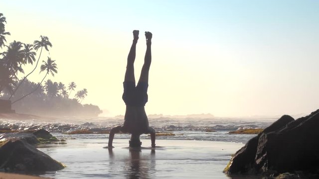 muscular athlete stands on hands and head against foaming ocean waves with brown rock on foreground extreme slow motion. Concept yoga and spiritual practices