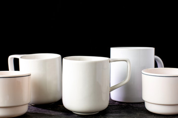 White tea and coffee mugs of various shapes on a wooden table and on a black background.