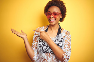 Young african american woman with afro hair wearing bikini and heart shaped sunglasses amazed and smiling to the camera while presenting with hand and pointing with finger.