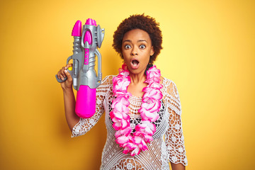 Young african american woman with afro hair wearing flower hawaiian lei and water gun scared in...