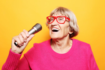 Portrait of funny grandmother holds up the microphone stand and sings isolated on vivid yellow...