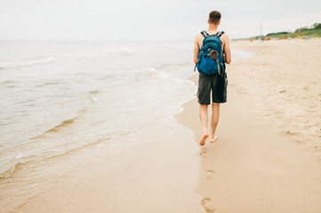 Young guy with backpack behind goes barefoot on beach rear view